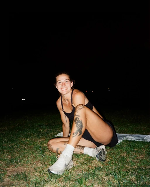Woman with tattoos sits on grass at night in athletic attire, smiling with white running shoes. Dark sky with subtle city lights.
