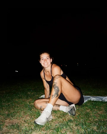 Woman with tattoos sits on grass at night in athletic attire, smiling with white running shoes. Dark sky with subtle city lights.