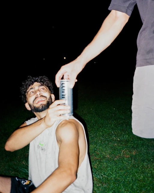 Two young adult men in casual clothing outside at night, one offering a can of sparkling water to the other on a lawn.
