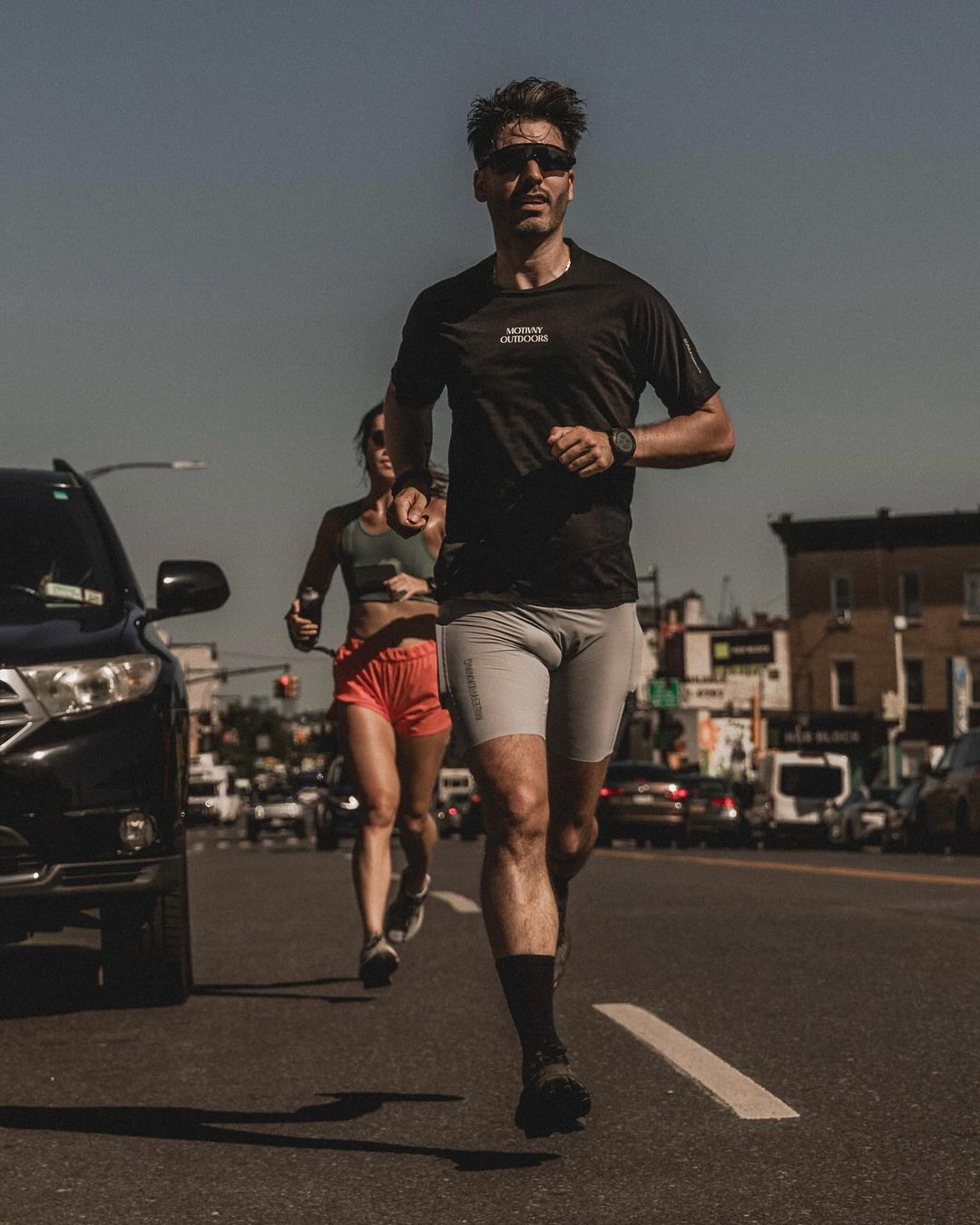 Man running down a city street wearing a black t-shirt and grey shorts with a woman running behind him