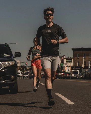 Man running down a city street wearing a black t-shirt and grey shorts with a woman running behind him