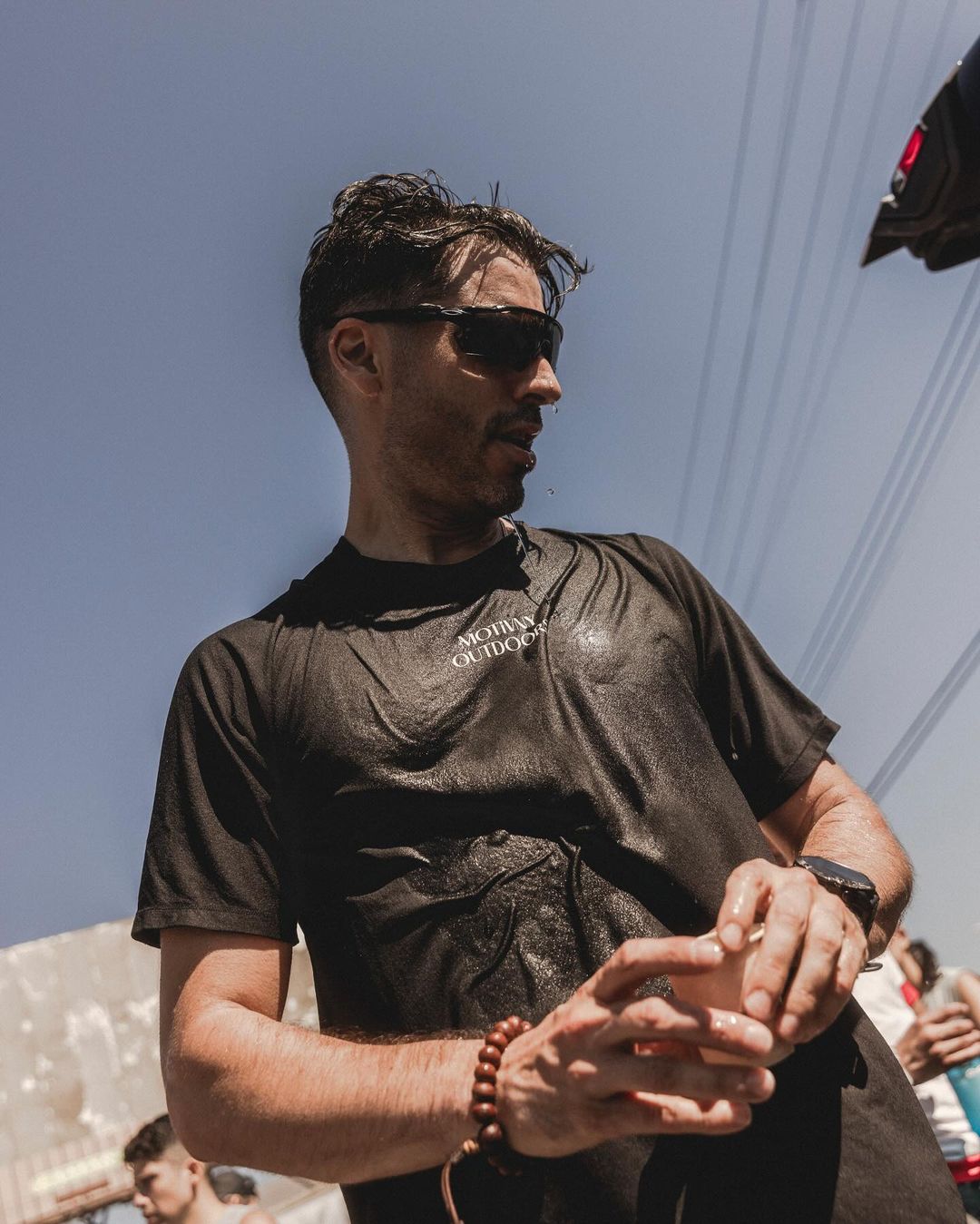Adult male with wet dark hair and sunglasses wearing a black t-shirt outdoors in natural light with electrical lines in the background.