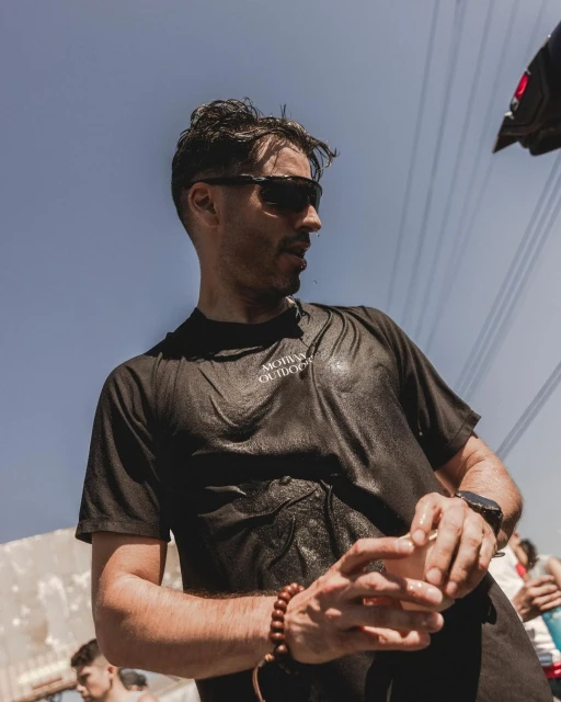 Adult male with wet dark hair and sunglasses wearing a black t-shirt outdoors in natural light with electrical lines in the background.