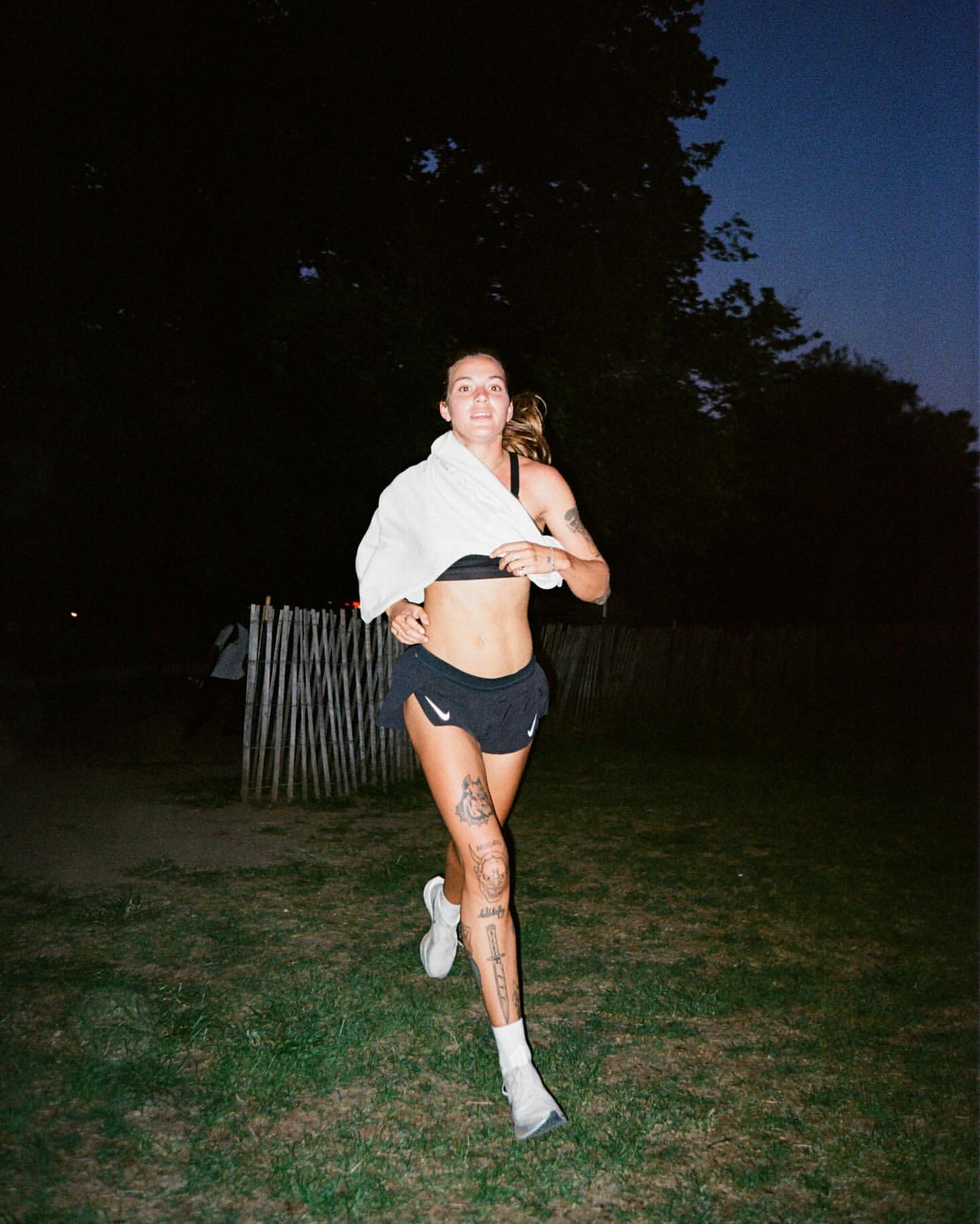 Adult female runner in athletic wear runs on a grassy field with a white towel draped over her shoulder at twilight.