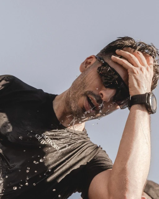 Athletic man douses face with water outdoors wearing sunglasses and black shirt, wrist watch visible against clear sky.