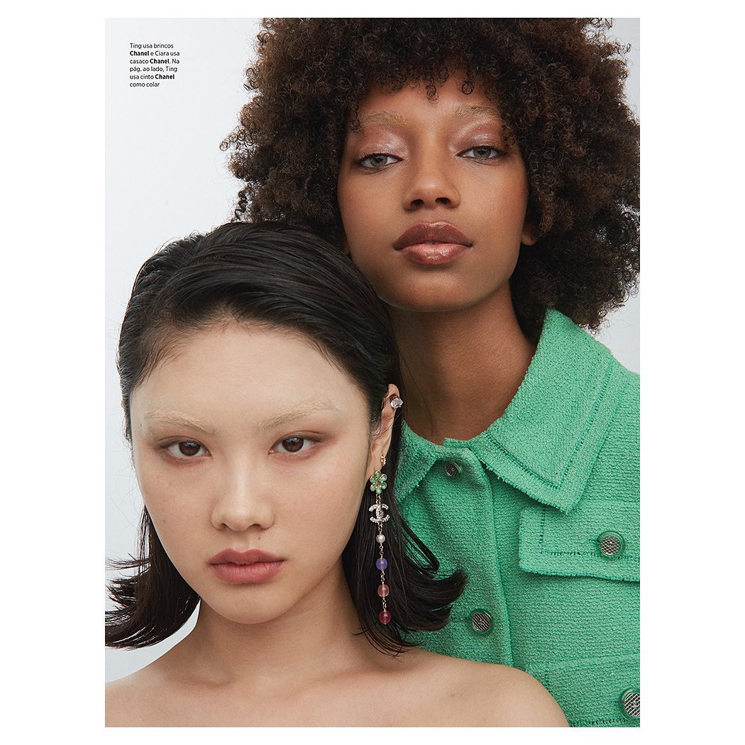 Close-up portrait of two female fashion models with glossy makeup, one in a green jacket and one wearing Chanel earrings in a studio setting.
