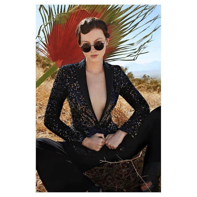 An adult female wearing a sequin jacket, sunglasses and black pants is seated in an outdoor desert landscape with a palm leaf.