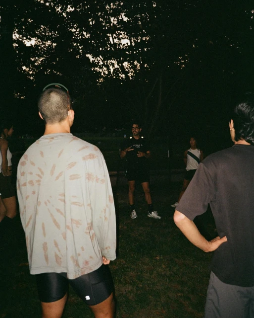 Group of people gathered outdoors at dusk; an adult male speaks to the group while wearing shorts, t-shirt and athletic shoes.
