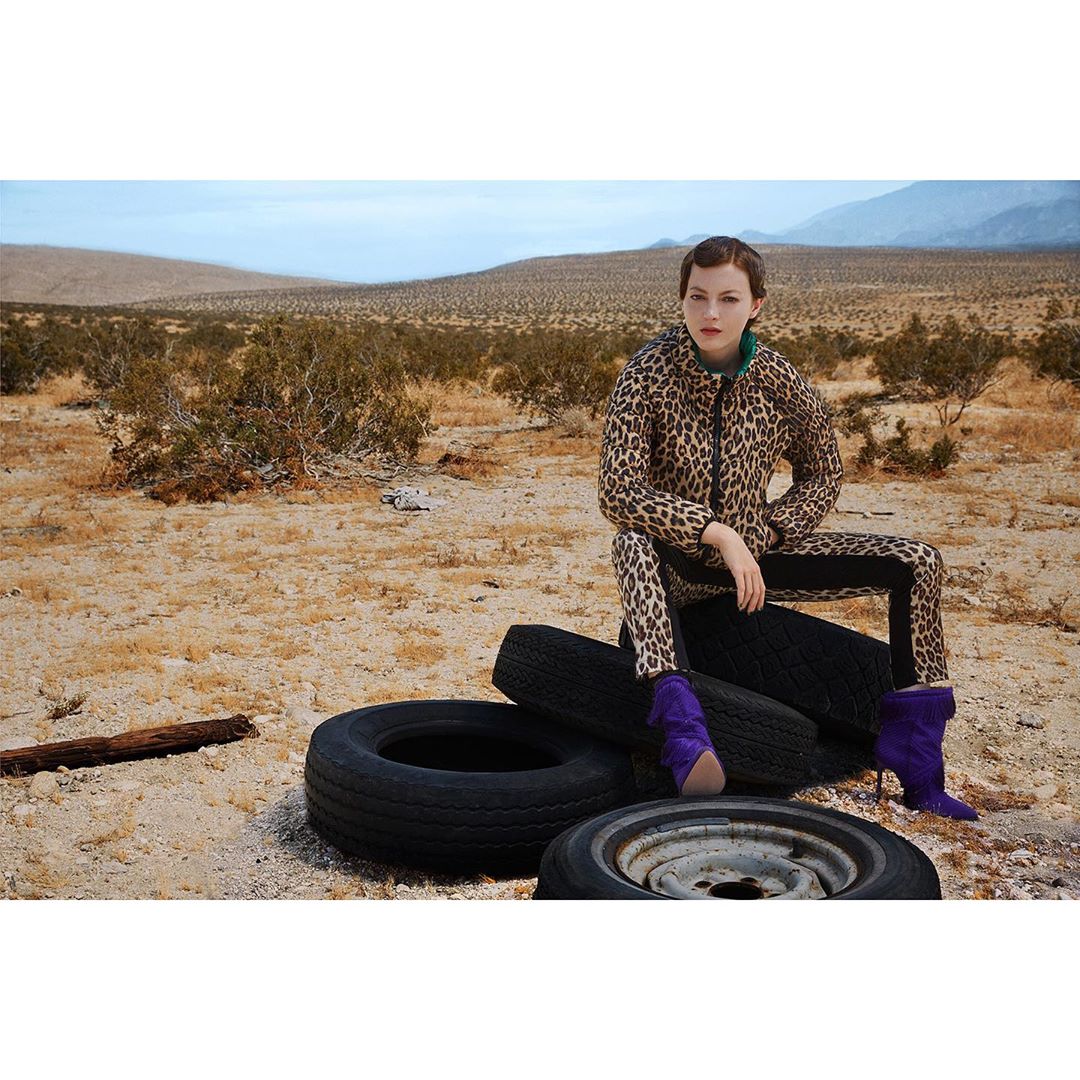 A woman in a leopard-print jacket and pants sits on a stack of tires in the desert wearing purple boots, creating a striking contrast.