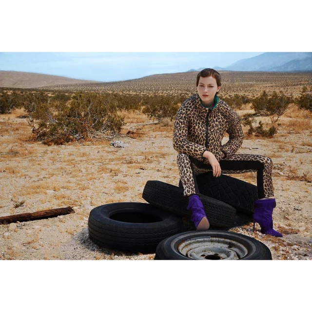 A woman in a leopard-print jacket and pants sits on a stack of tires in the desert wearing purple boots, creating a striking contrast.