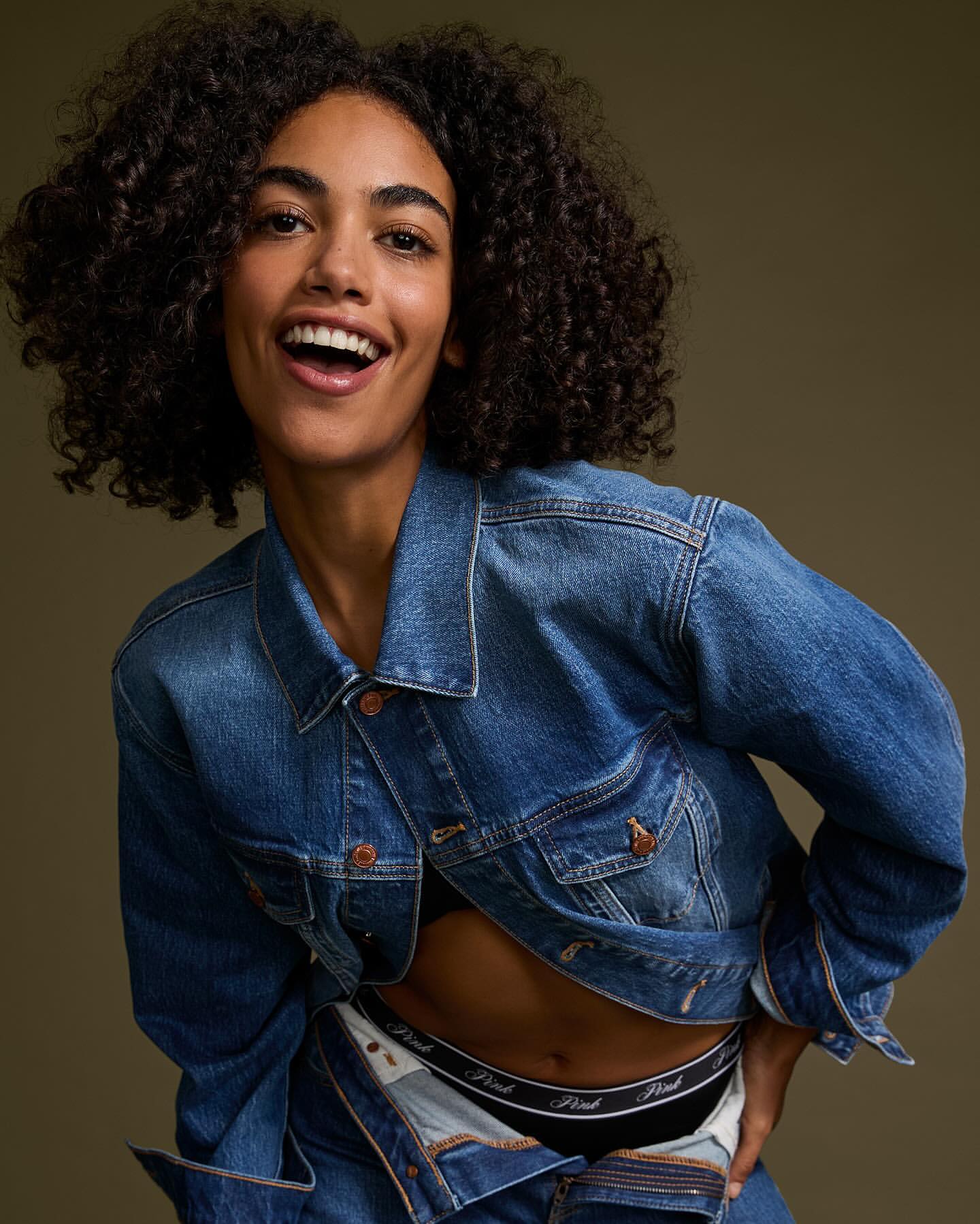 A smiling woman with curly hair wears a denim jacket, revealing a black bra and underwear, posing in a photo studio with a neutral background.