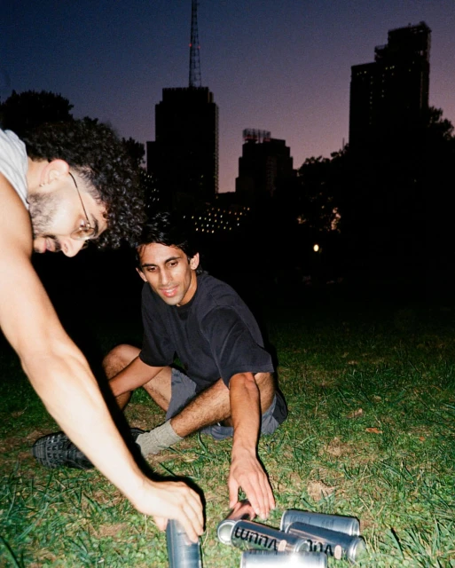 Two young men gather around a collection of spray paint cans on a grassy field at night in front of a city skyline.