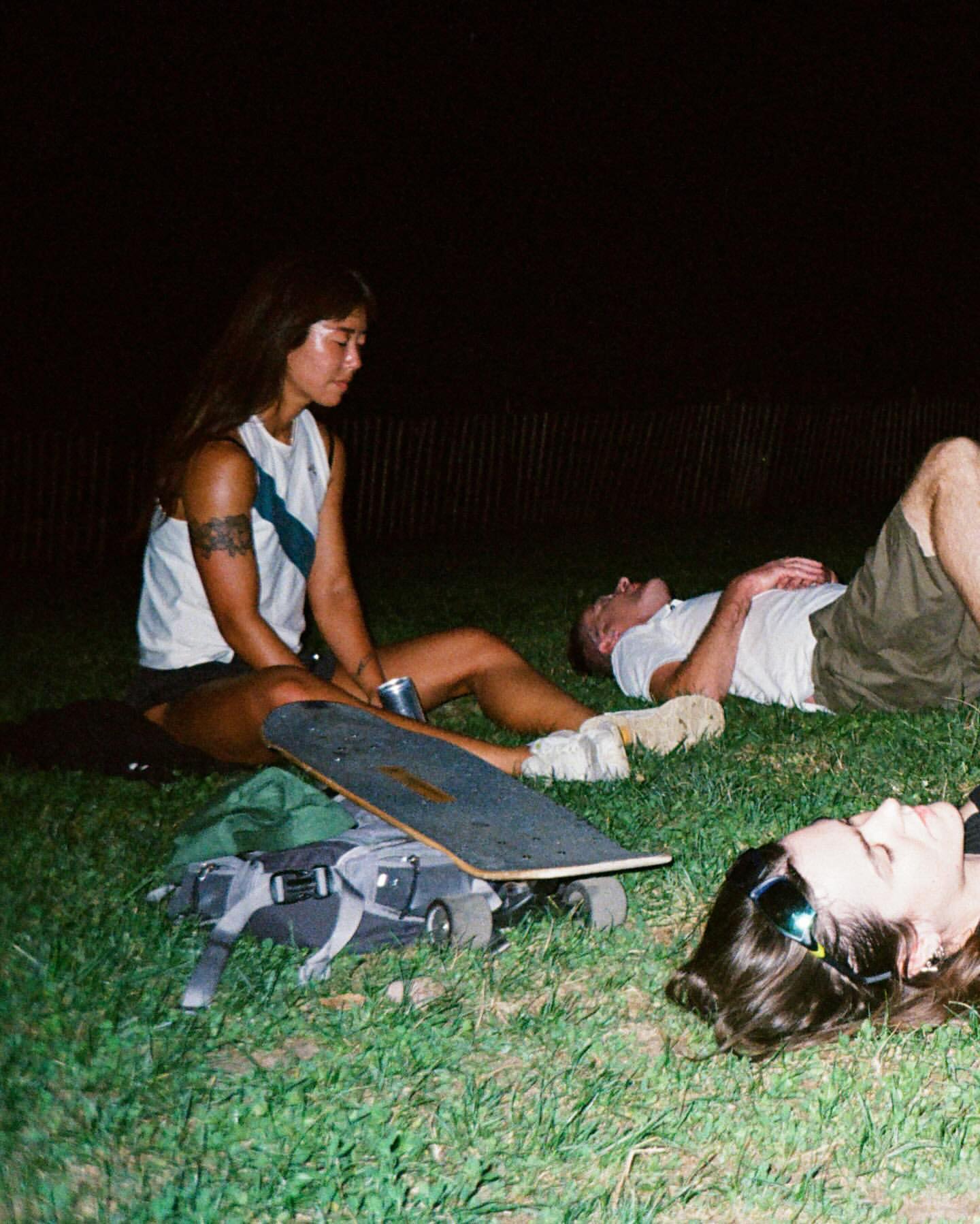 Three people relax on grass at night with a skateboard, backpack, and beverage; two are reclining, and one is sitting with long hair and tattoos.