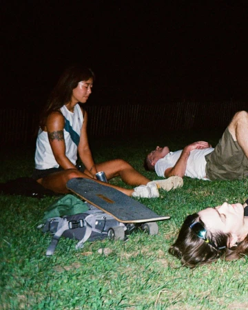 Three people relax on grass at night with a skateboard, backpack, and beverage; two are reclining, and one is sitting with long hair and tattoos.