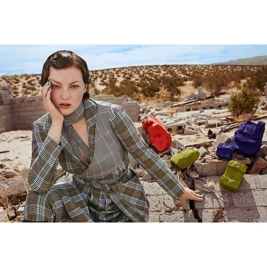 A young woman poses in a plaid suit and scarf amidst a desert landscape with crumbling structures and colorful painted blocks.