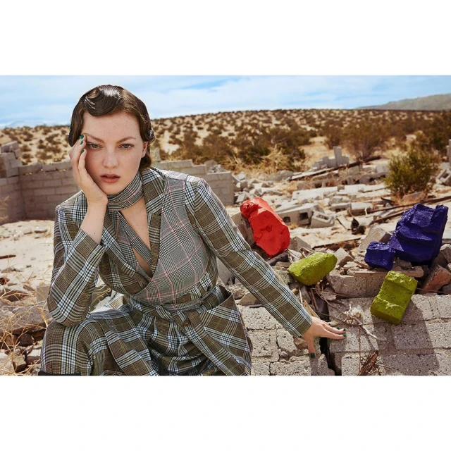 A young woman poses in a plaid suit and scarf amidst a desert landscape with crumbling structures and colorful painted blocks.