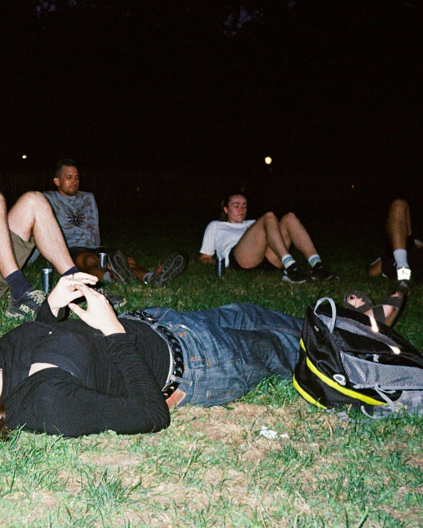 Group of adults relaxing on grass at night, casual clothing, drinks visible, dark ambiance, leisure, outdoor gathering, friendship.