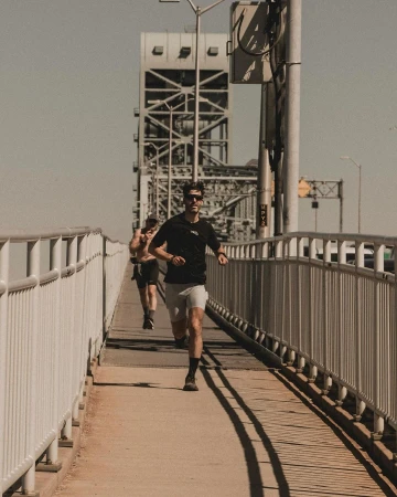 Man running across a metal bridge pathway wearing dark t-shirt and sunglasses, photographed by Aingeru Zorita.