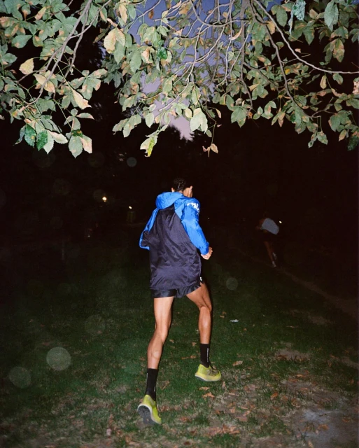 A man in running attire jogs through a grassy field at night, illuminated by soft light filtering through lush tree branches overhead.