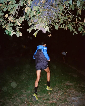 A man in running attire jogs through a grassy field at night, illuminated by soft light filtering through lush tree branches overhead.