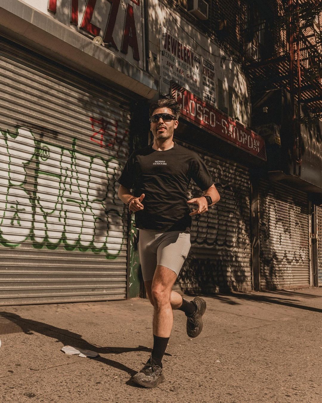Man running on a city street wearing sportswear, t-shirt, and sunglasses, with graffiti-covered storefronts and urban buildings in the background.