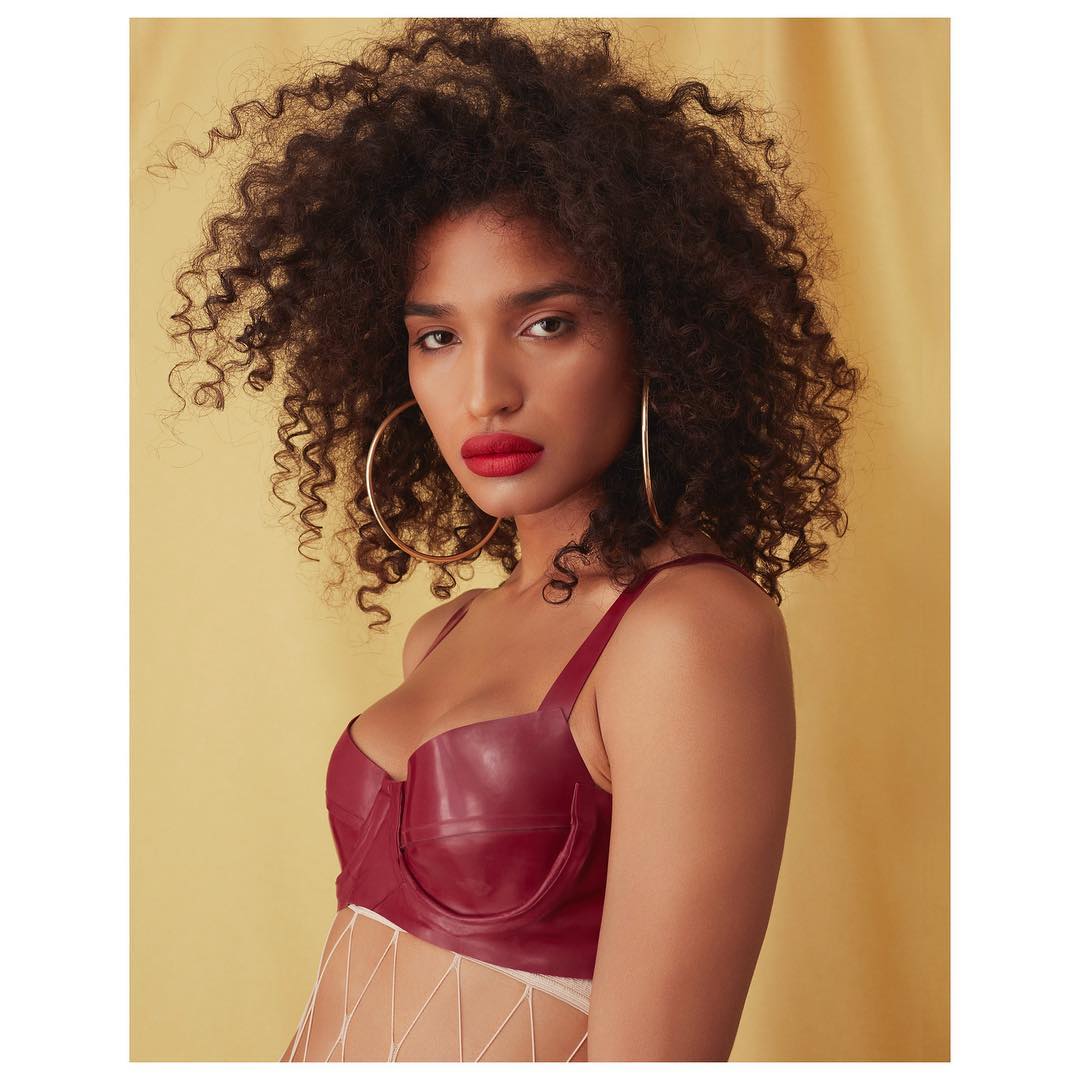 Portrait of a young woman with curly hair, wearing a red top and gold hoop earrings against a yellow background in a photo studio.