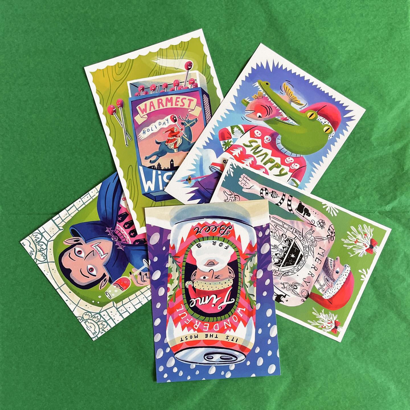 A collection of illustrated greeting cards featuring various cartoonish holiday and Christmas themed designs are arranged on green tissue paper.
