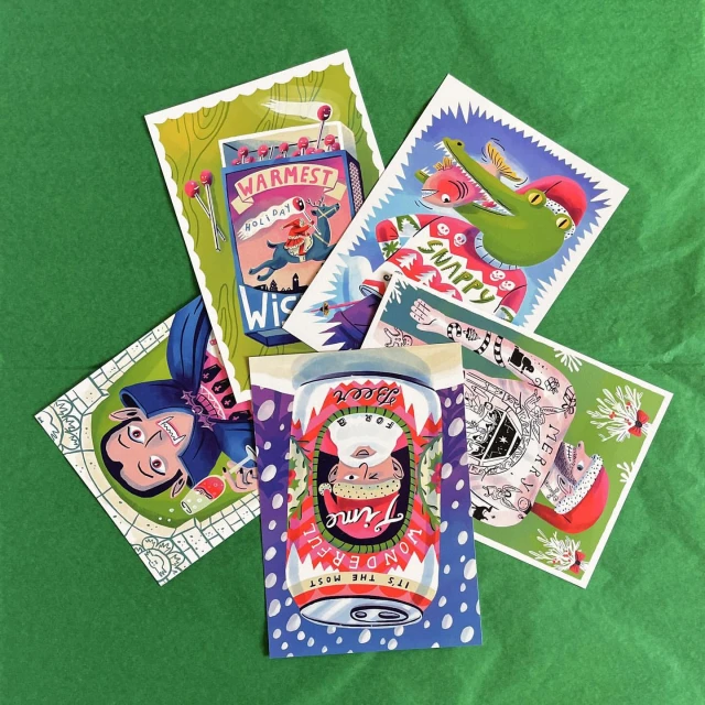A collection of illustrated greeting cards featuring various cartoonish holiday and Christmas themed designs are arranged on green tissue paper.