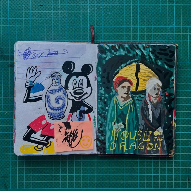 Handmade sketchbook displays cartoon characters and painting of 'House of the Dragon' figures, set on a green cutting mat.