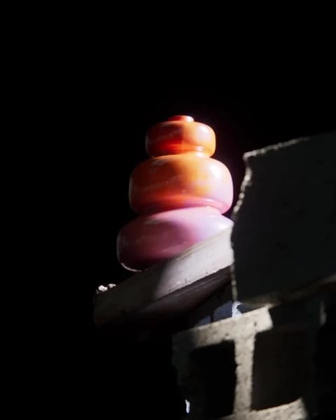 Abstract still life of stacked colorful glass-like forms in the dark against a rough, gray concrete block wall with shadows.