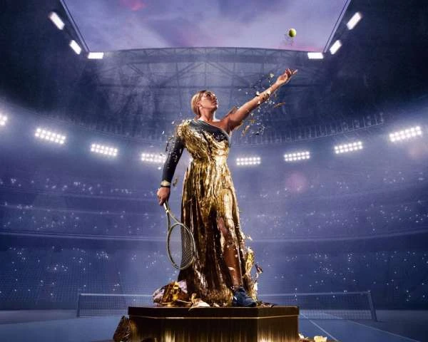 Female tennis player in a gold gown serves a tennis ball in an indoor stadium with bright lights and a large audience in the background.