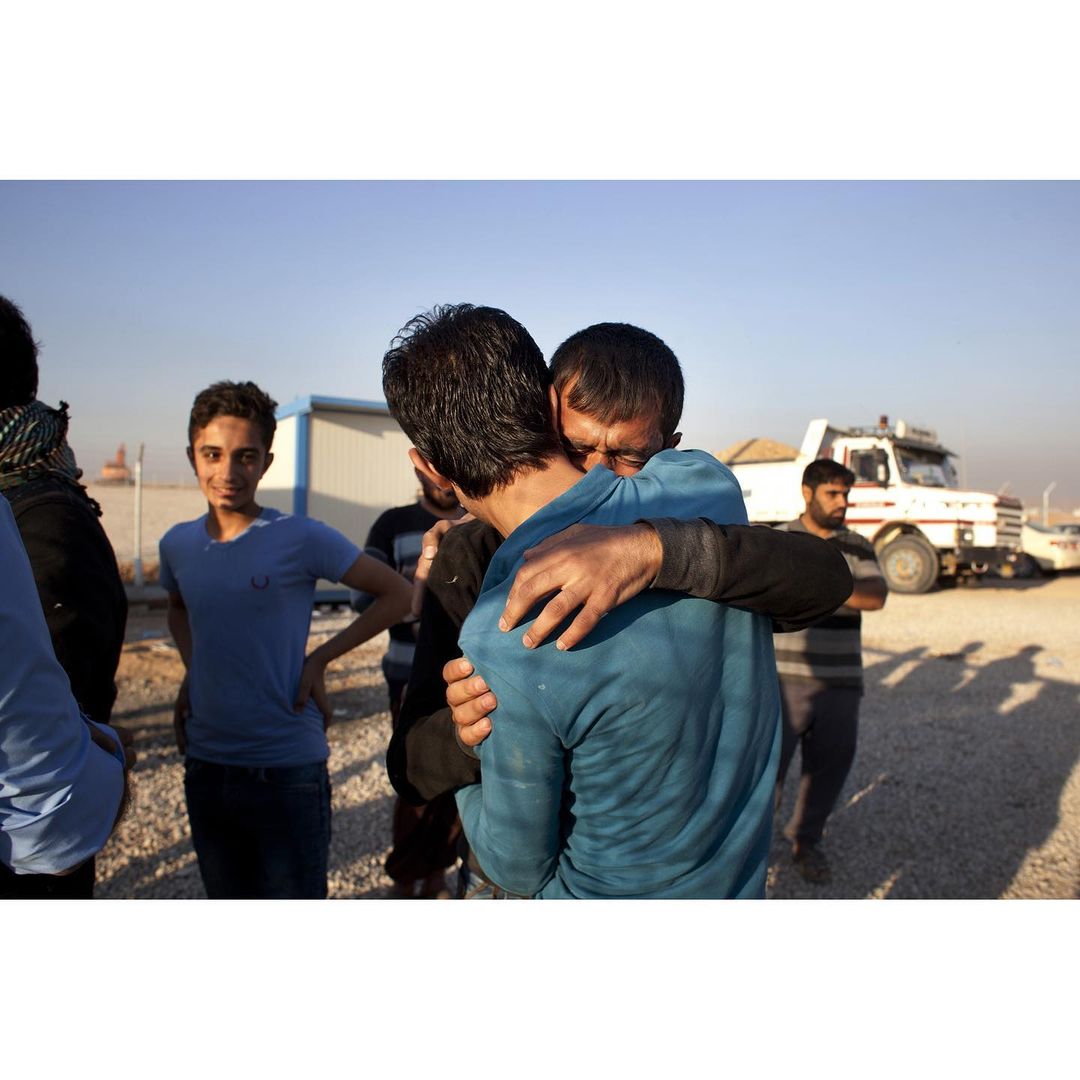 Two adult men embrace warmly outdoors, with a group of people and a utility vehicle in the background on a sunny day.