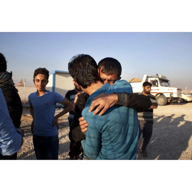 Two adult men embrace warmly outdoors, with a group of people and a utility vehicle in the background on a sunny day.