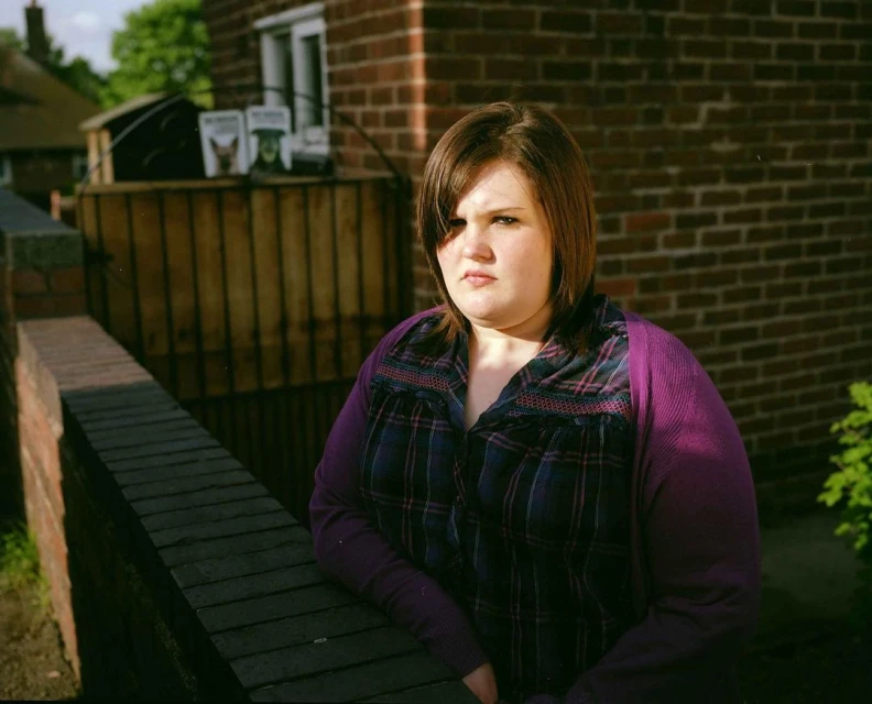 A young woman with short brown hair poses outdoors near a brick wall wearing a plaid shirt and purple cardigan in natural light.
