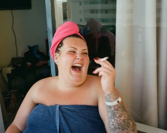 Smiling woman wearing a pink towel on her head and a blue towel wrap, holding a lit cigarette.