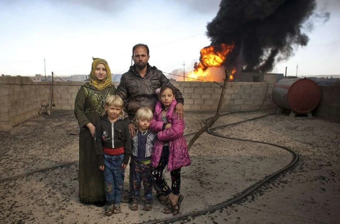 Family of five stands in front of billowing smoke and fire; mother in hijab, father with arm on daughter, two small blonde children.
