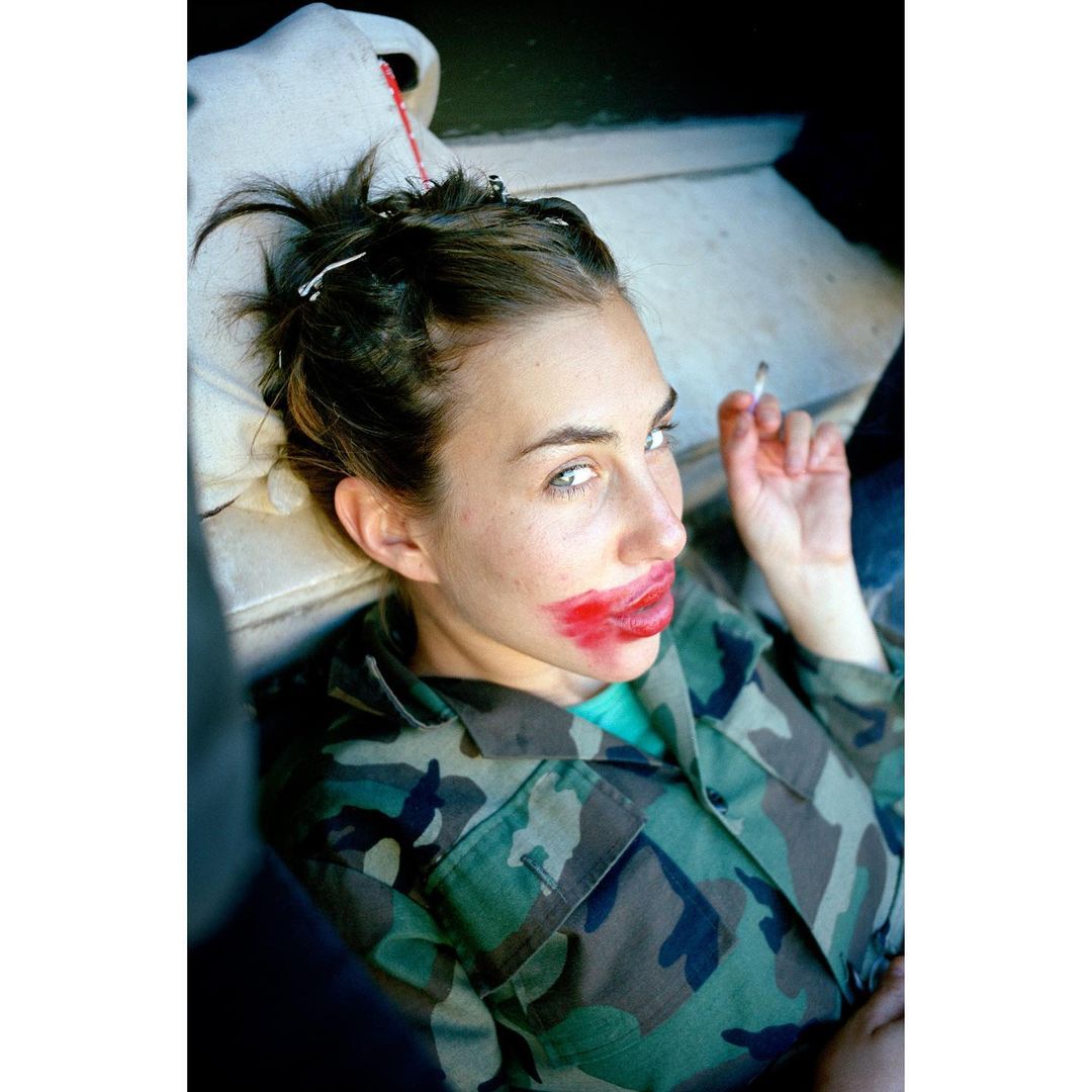 Woman with smeared lipstick, lying down in a camo shirt, holds a cigarette, resting on a stone surface, portrait-style image.