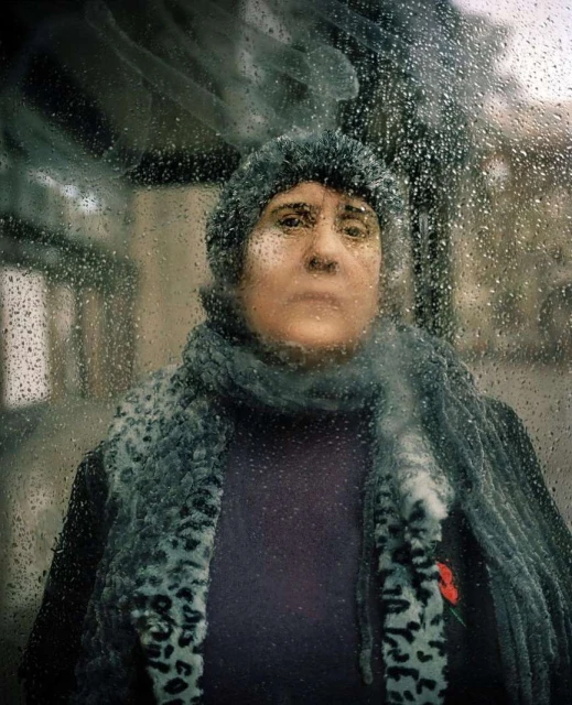Portrait of a mature woman with knit hat and scarf, seen through a rain-streaked window, creating a blurred and reflective effect.