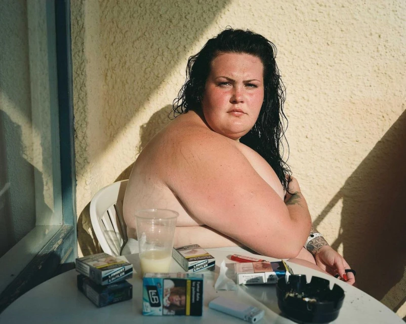 Overweight woman sits at a table with cigarettes and a drink outside, facing the camera in natural light. Neutral expression.