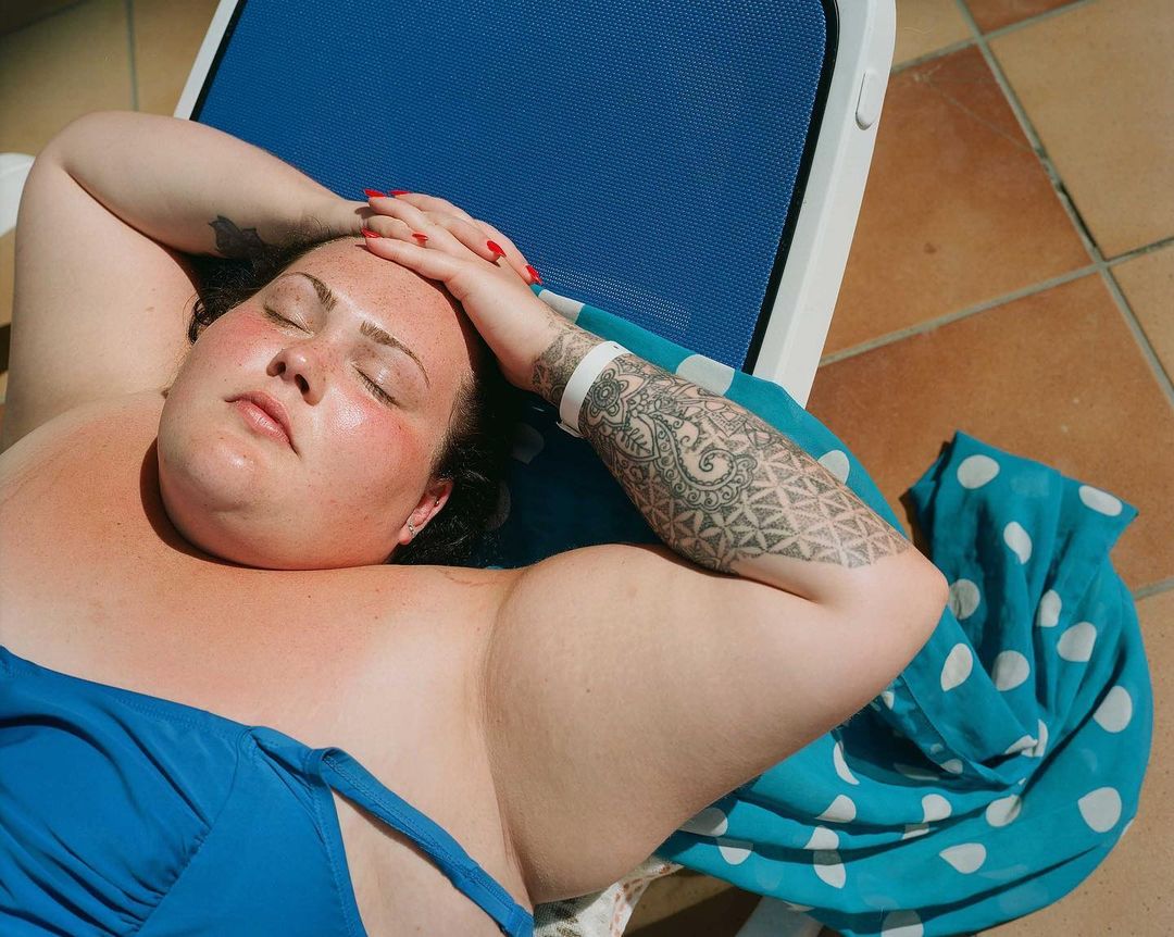 Adult woman relaxing on a lounge chair outdoors in the sun, wearing a blue bathing suit, with a patterned cover and tile flooring.