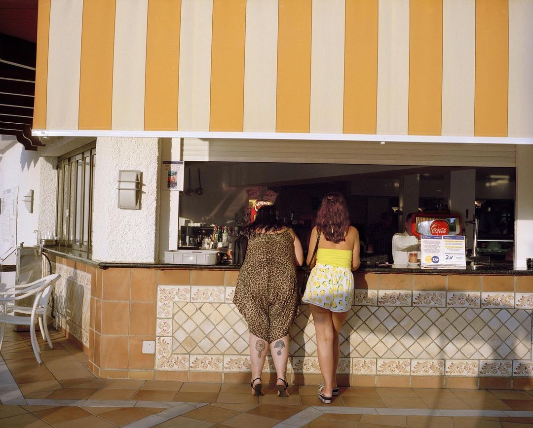 Two women stand at a bar on a sunny outdoor patio with orange and white striped awning above, drinks and bottles visible.