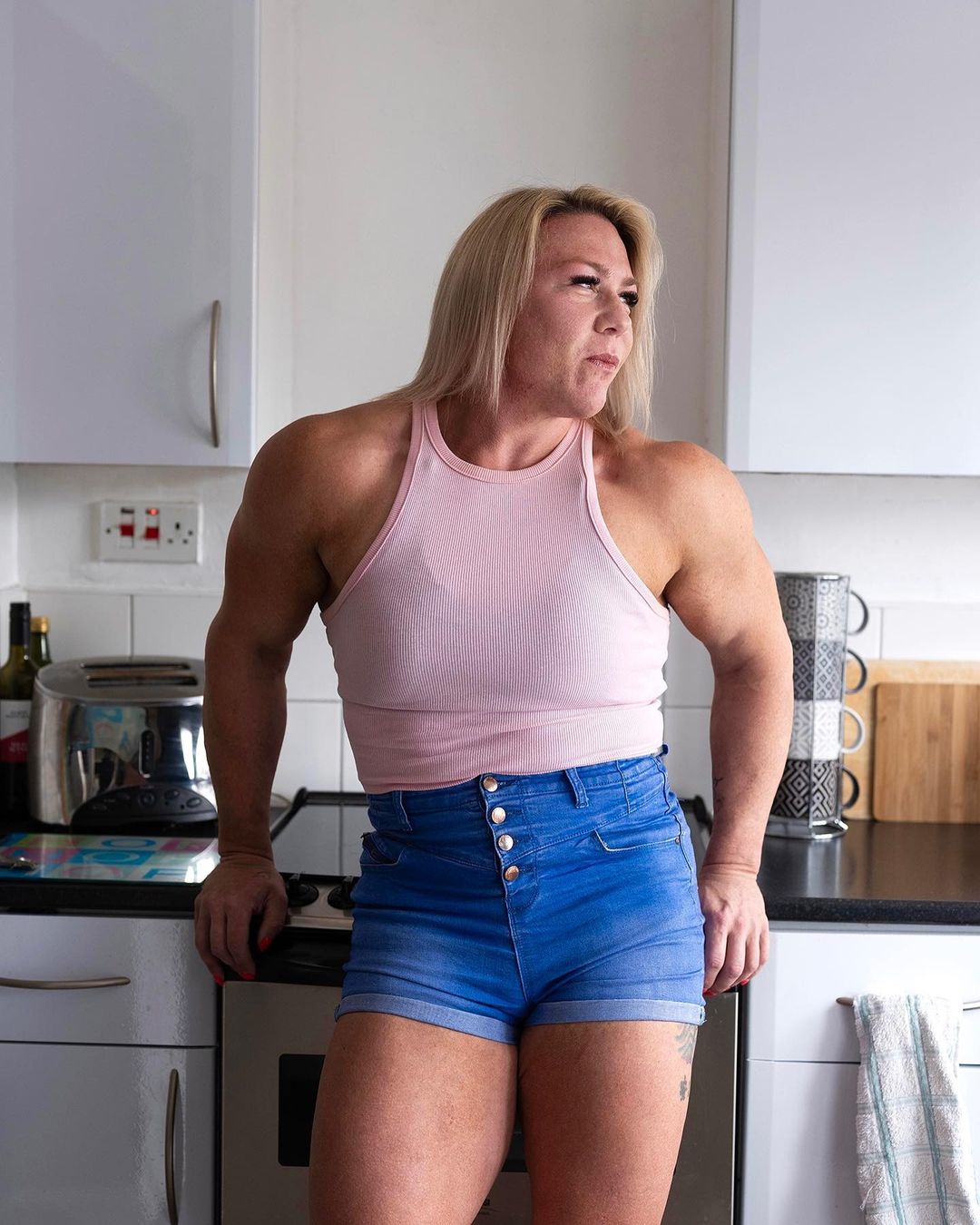 Muscular woman in denim shorts and a pink tank top poses indoors near a kitchen counter with a stoic expression and light skin.