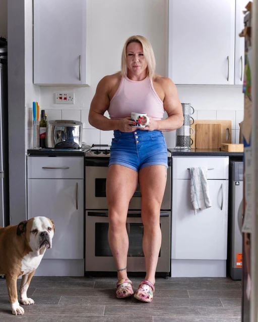 Muscular woman in tank top and jean shorts holds mug in a kitchen with a bulldog, capturing a fitness and lifestyle moment.