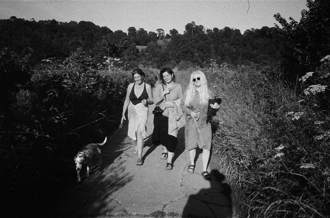 Three adult women walk a dog along a path lined with tall grass and greenery, backed by a forest in a black and white photograph.
