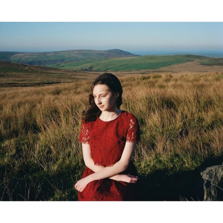 Young woman in a red lace dress seated outdoors amidst a field of tall dry grass under an expansive landscape with rolling hills.