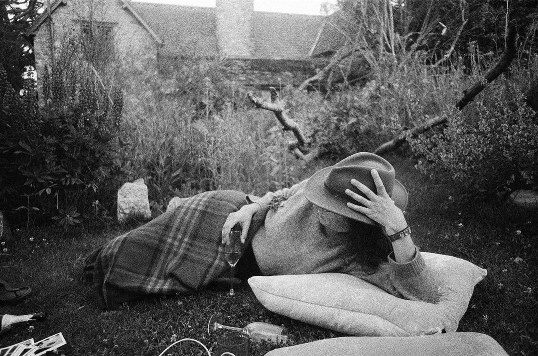 Woman in hat lies on pillow in backyard, relaxing with wine. Picnic setting, plaid blanket, house in background, black and white.