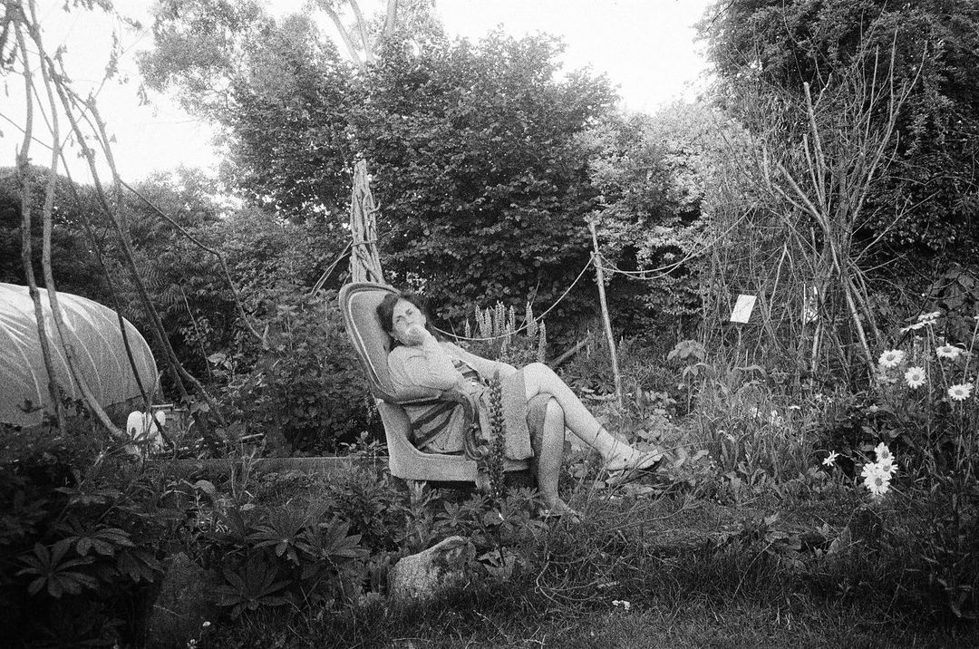 Black and white image of a woman relaxing in a chair outdoors surrounded by greenery and daisies. Soft lighting. 