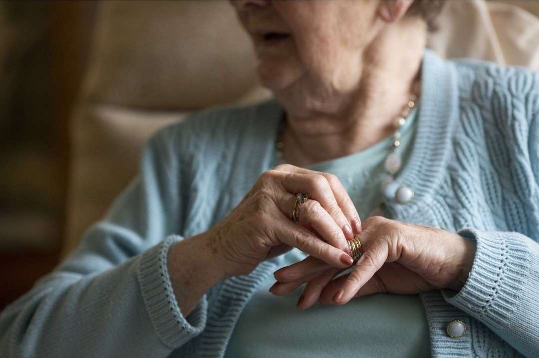 Elderly woman sitting indoors wearing a light blue cardigan, with her hands adjusting rings on her fingers, captured in soft, natural light.