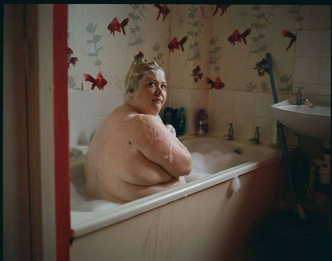 Woman with blonde hair is sitting in a white bathtub with bubbles, looking towards the viewer in an indoor bathroom setting.