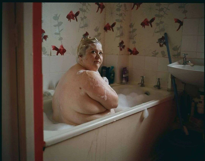 Woman with blonde hair is sitting in a white bathtub with bubbles, looking towards the viewer in an indoor bathroom setting.